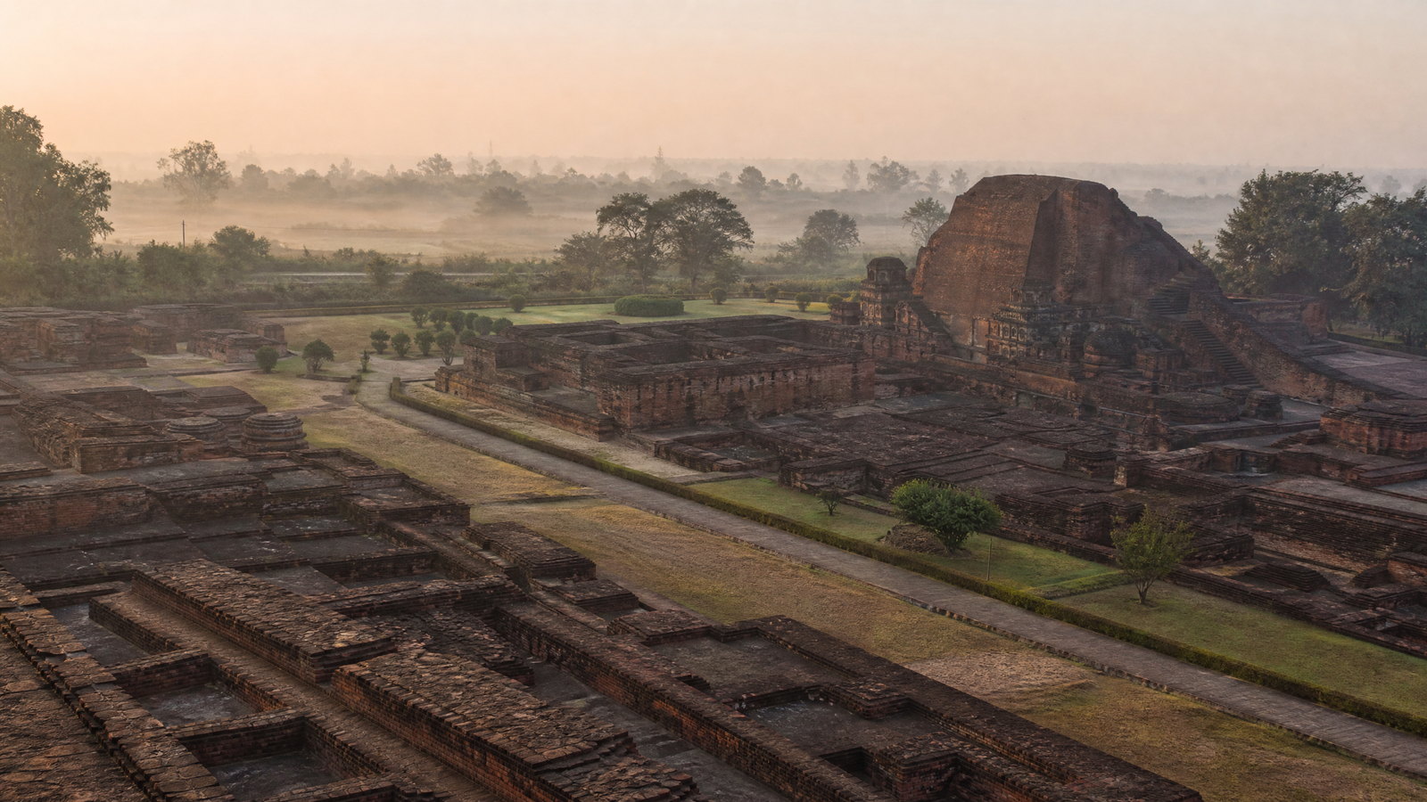 Early light over the red-brick terraces and votive shrines of Nalanda Mahavihara, with low winter mist above surrounding fields.