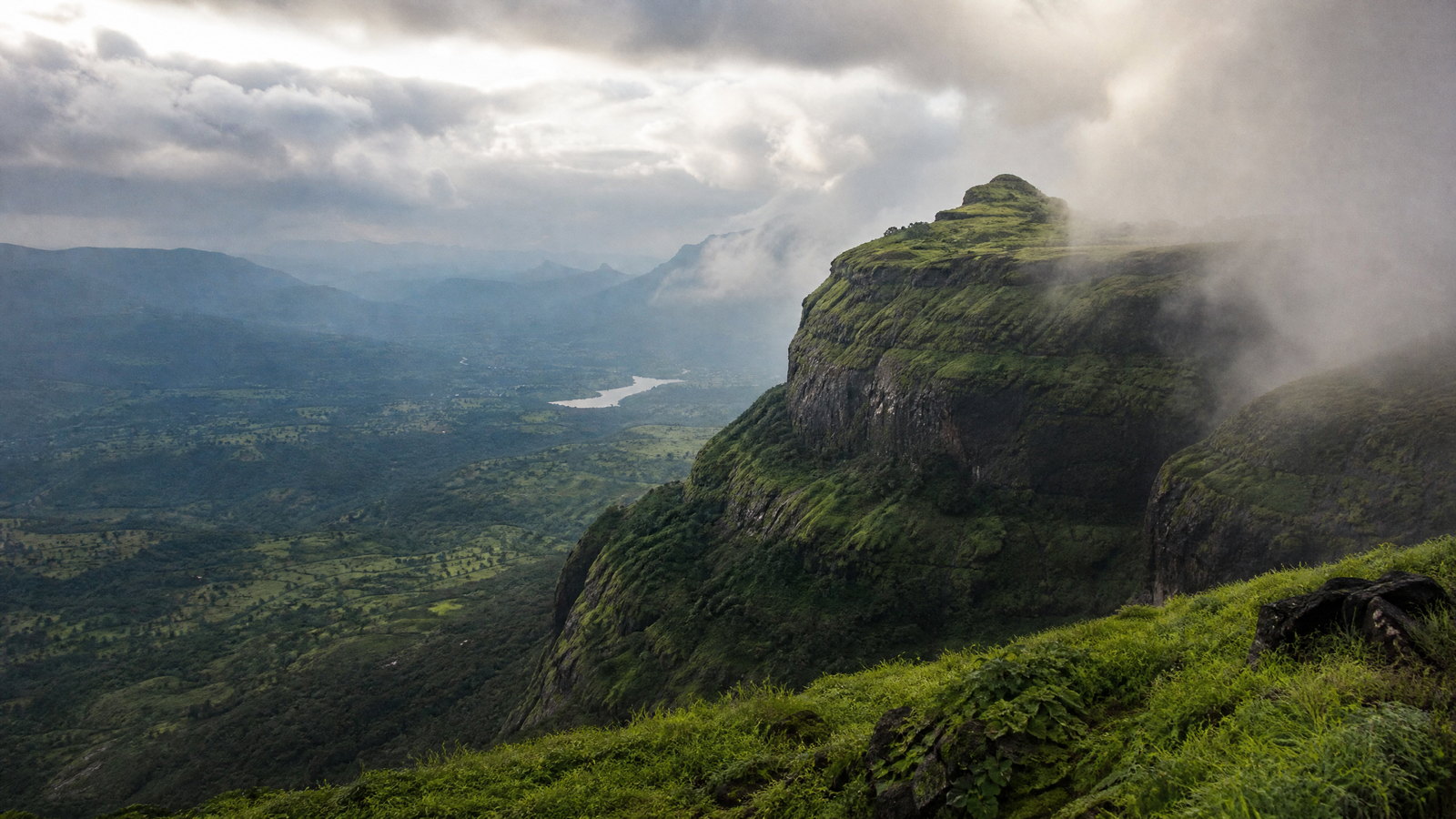 Monsoon clouds gathering over the layered basalt escarpment and green plateau of the Sahyadri hills in Maharashtra.