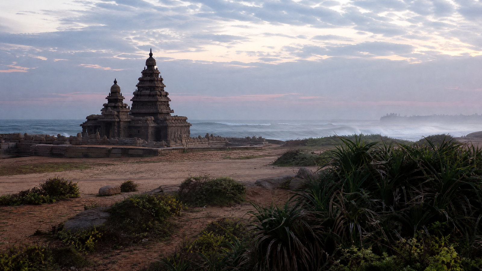 Granite Shore Temple at Mamallapuram standing above the Bay of Bengal at first light, with empty sand and a pale horizon.