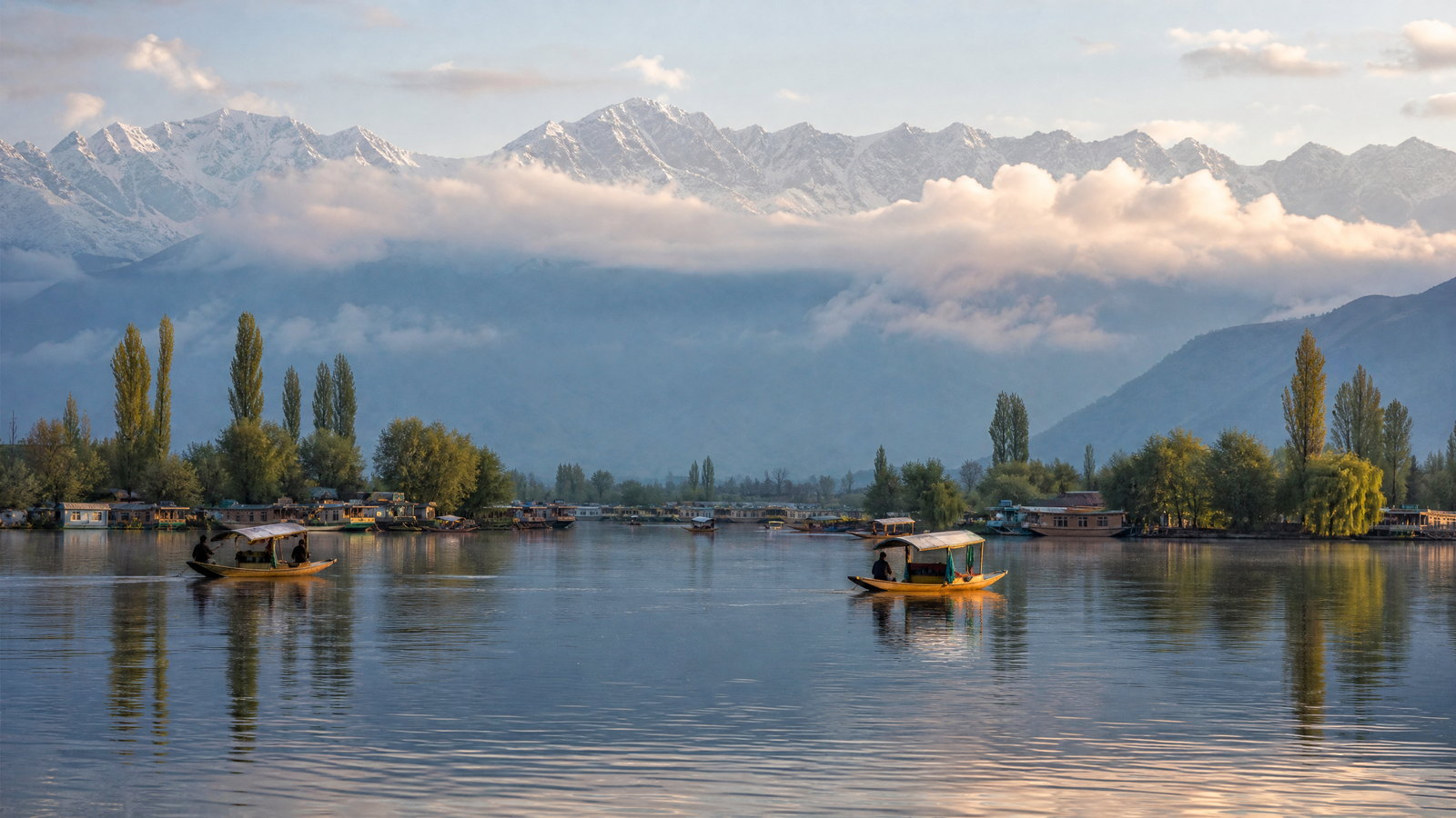A quiet early-morning view across Dal Lake toward Srinagar’s wooded Zabarwan foothills, with traditional wooden boats moored beside reed-lined water and snow-dusted mountains beyond.