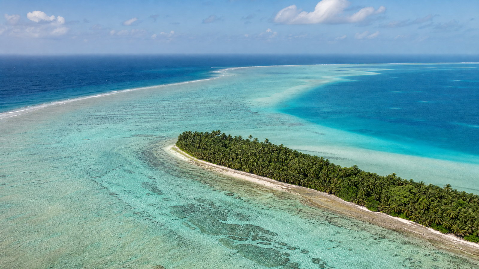 Aerial view of a narrow coconut-covered Lakshadweep coral island curving around a pale turquoise lagoon and bright reef edge.