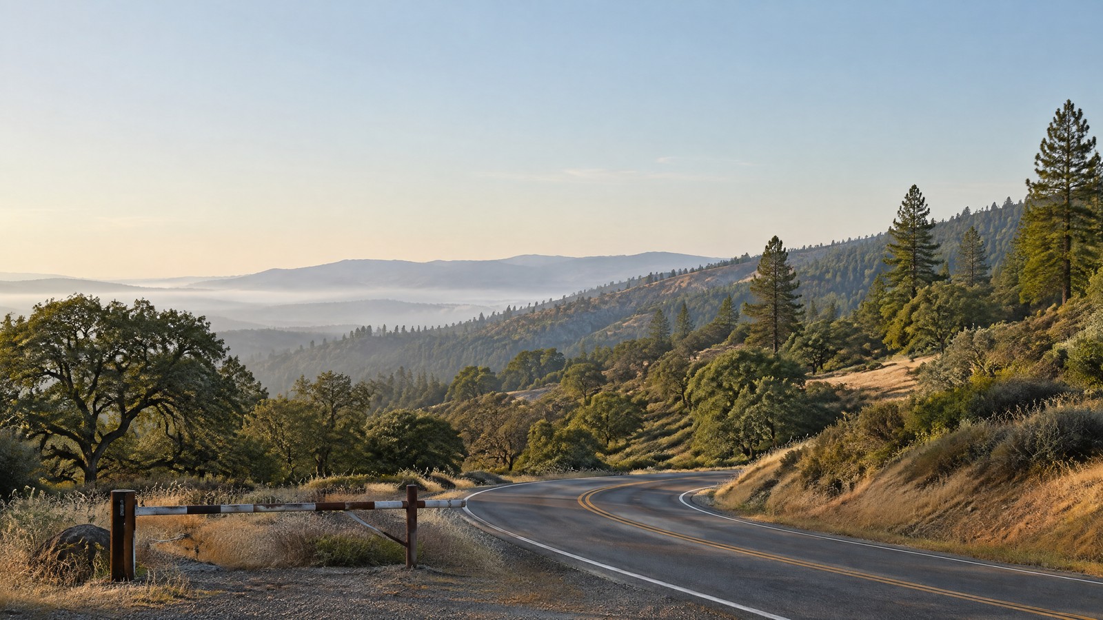 A calm northern California oak-and-pine ridge at early morning, with a two-lane rural road and a small unbranded road-control barrier in the foreground; pale haze sits far beyond the road.